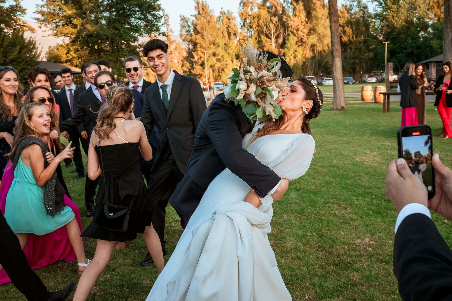 Groom dips bride joyfully surrounded by celebrating wedding guests on green lawn at outdoor reception.