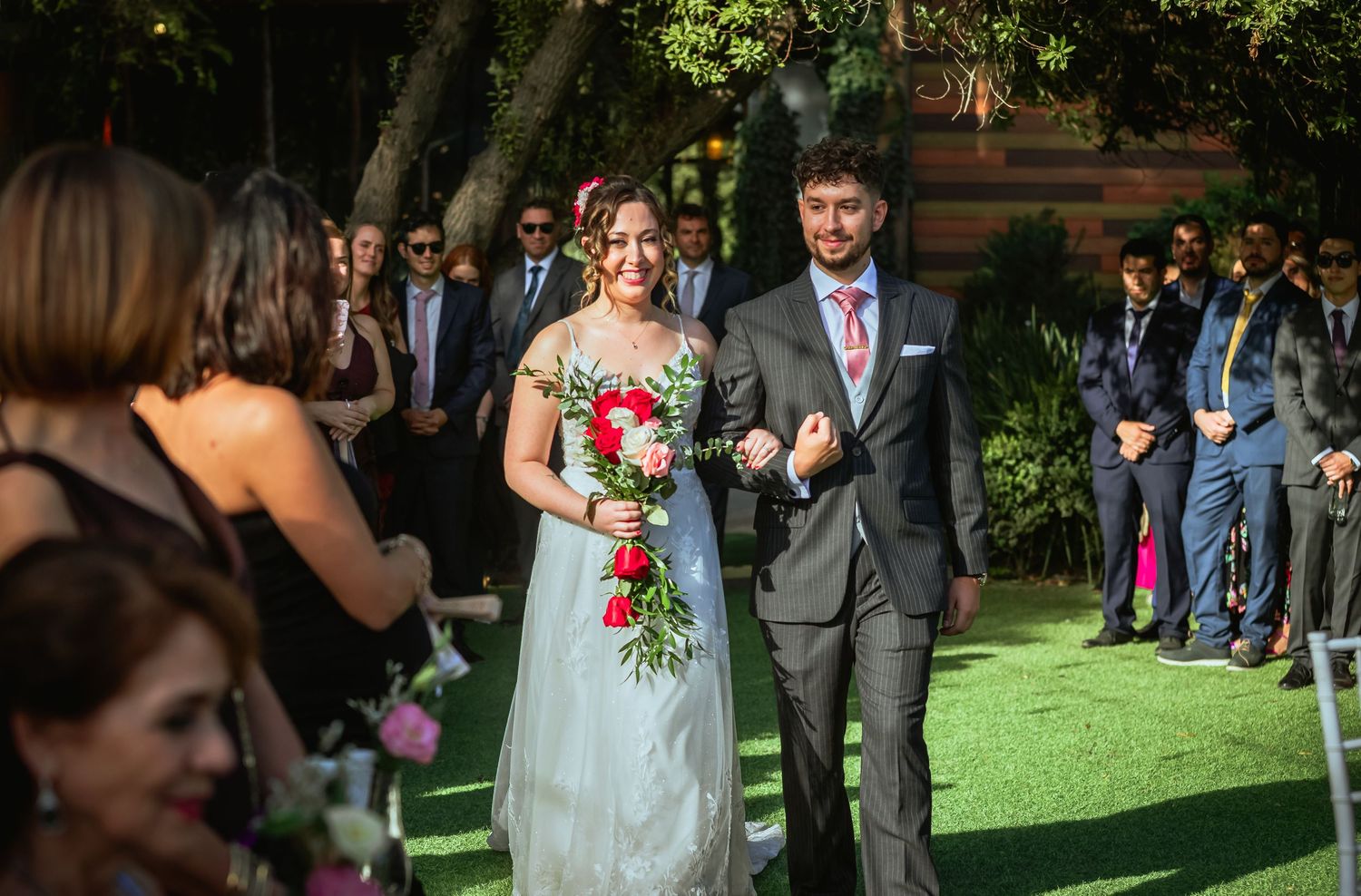 Bride and groom walk down outdoor aisle smiling, bride holding red bouquet at garden wedding.