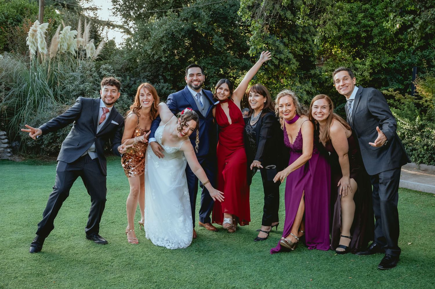 Bride and groom pose with smiling wedding guests in colorful attire at outdoor garden celebration.