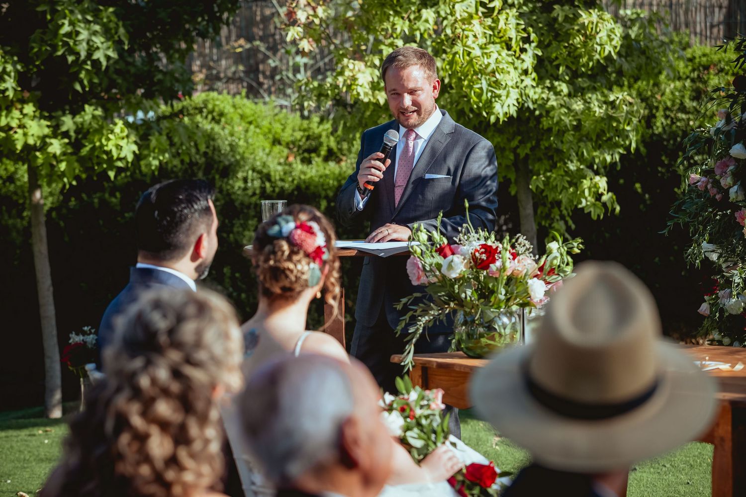 Officiant in dark suit speaks into microphone before bride and groom at outdoor garden wedding.