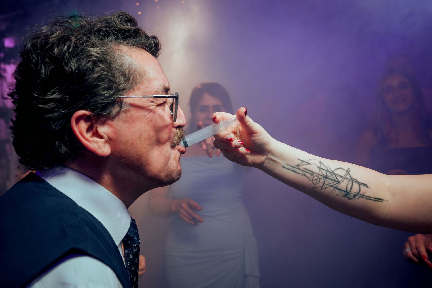 Man in vest smoking at lively party with purple smoke and tattooed hand offering cigarette.