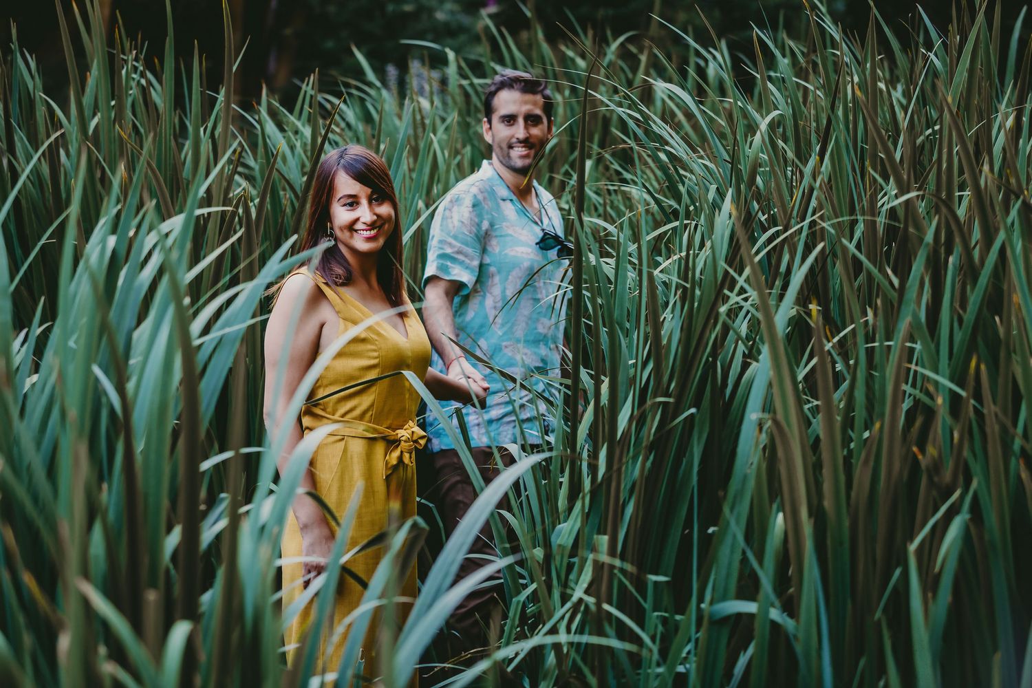 Happy couple walking hand in hand through tall green reeds in a lush garden setting.