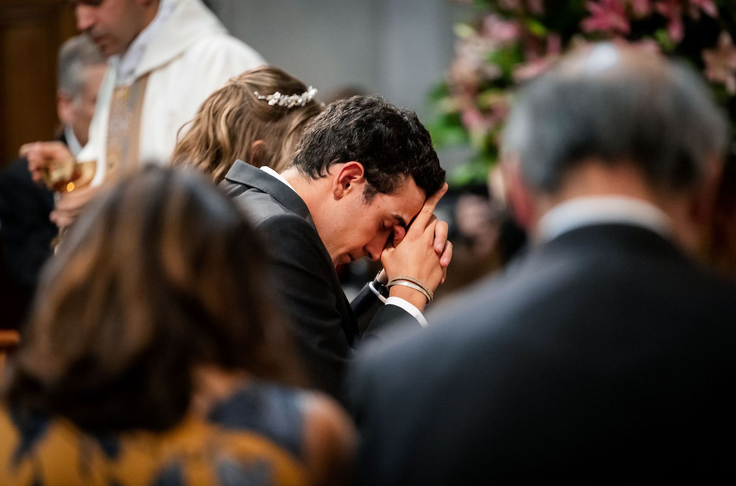 Man in suit bowing head in prayer during a church wedding ceremony with guests around him.
