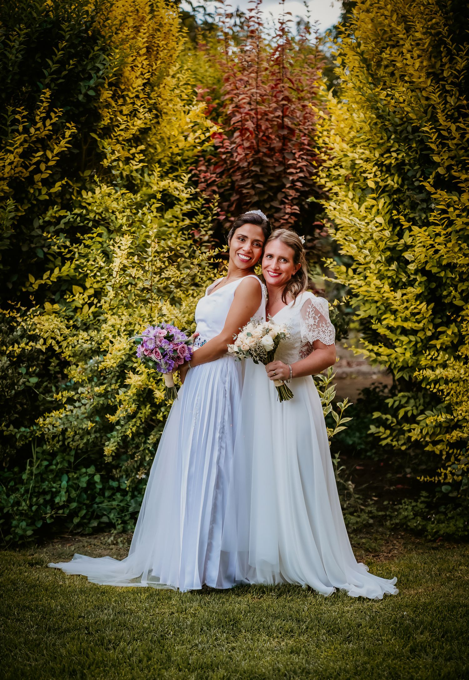 Two brides in white gowns holding bouquets pose together in a lush garden with colorful foliage.