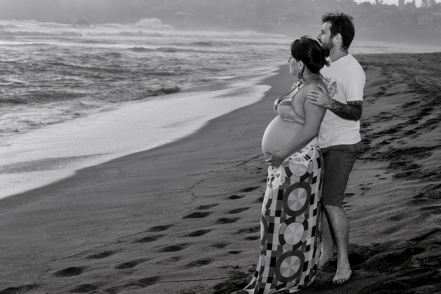 Pregnant couple embracing on a beach shoreline in black and white maternity photography.