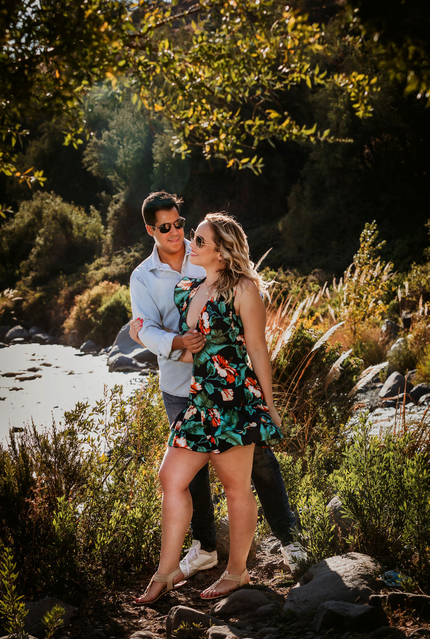 Couple embracing outdoors near a river, woman in floral dress and man in light blue shirt, surrounded by lush greenery.