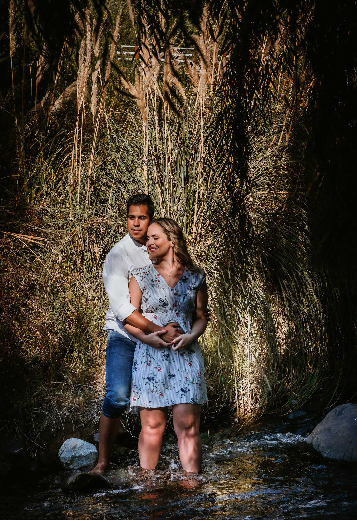 Couple standing in a shallow creek surrounded by tall grass, embracing tenderly in a romantic outdoor maternity photo shoot.