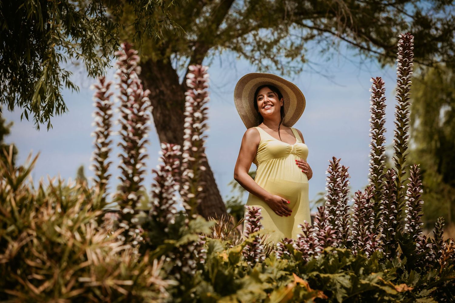 Pregnant woman in yellow dress and sun hat standing among purple flowers in a sunny garden.