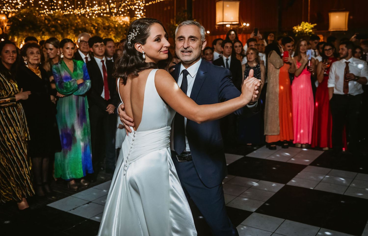 Bride and groom share first dance on checkered floor at elegant wedding reception with guests watching.