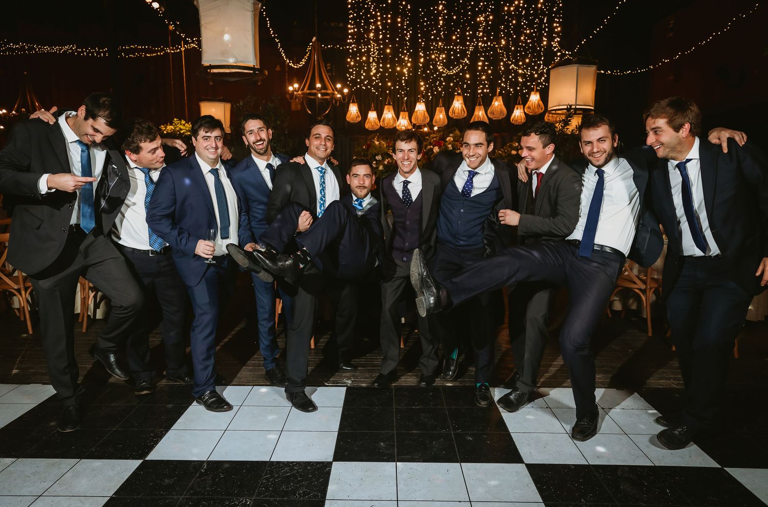 Groomsmen in navy suits dancing and posing on a black and white checkered dance floor at a wedding reception.
