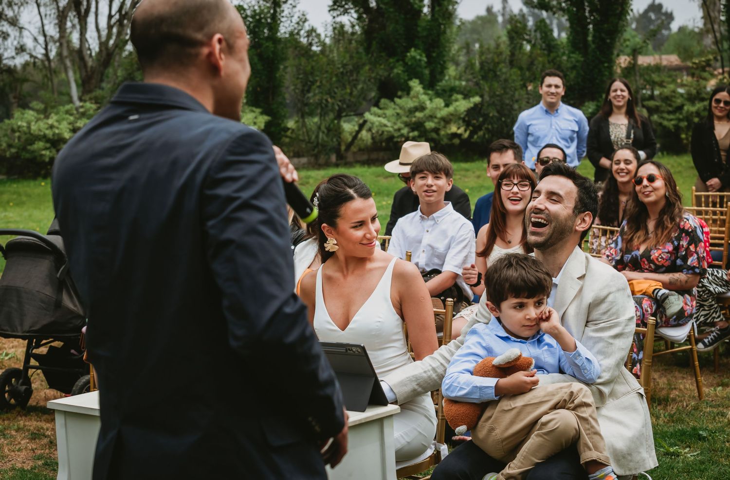 Joyful wedding guests laughing during an outdoor ceremony, with a couple holding a child seated in white chairs.