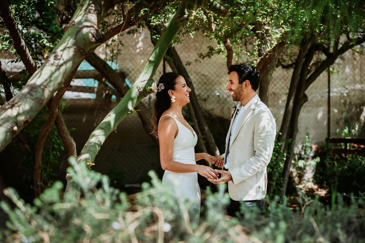 Couple exchanging vows outdoors under shaded trees, bride in white dress and groom in white suit.