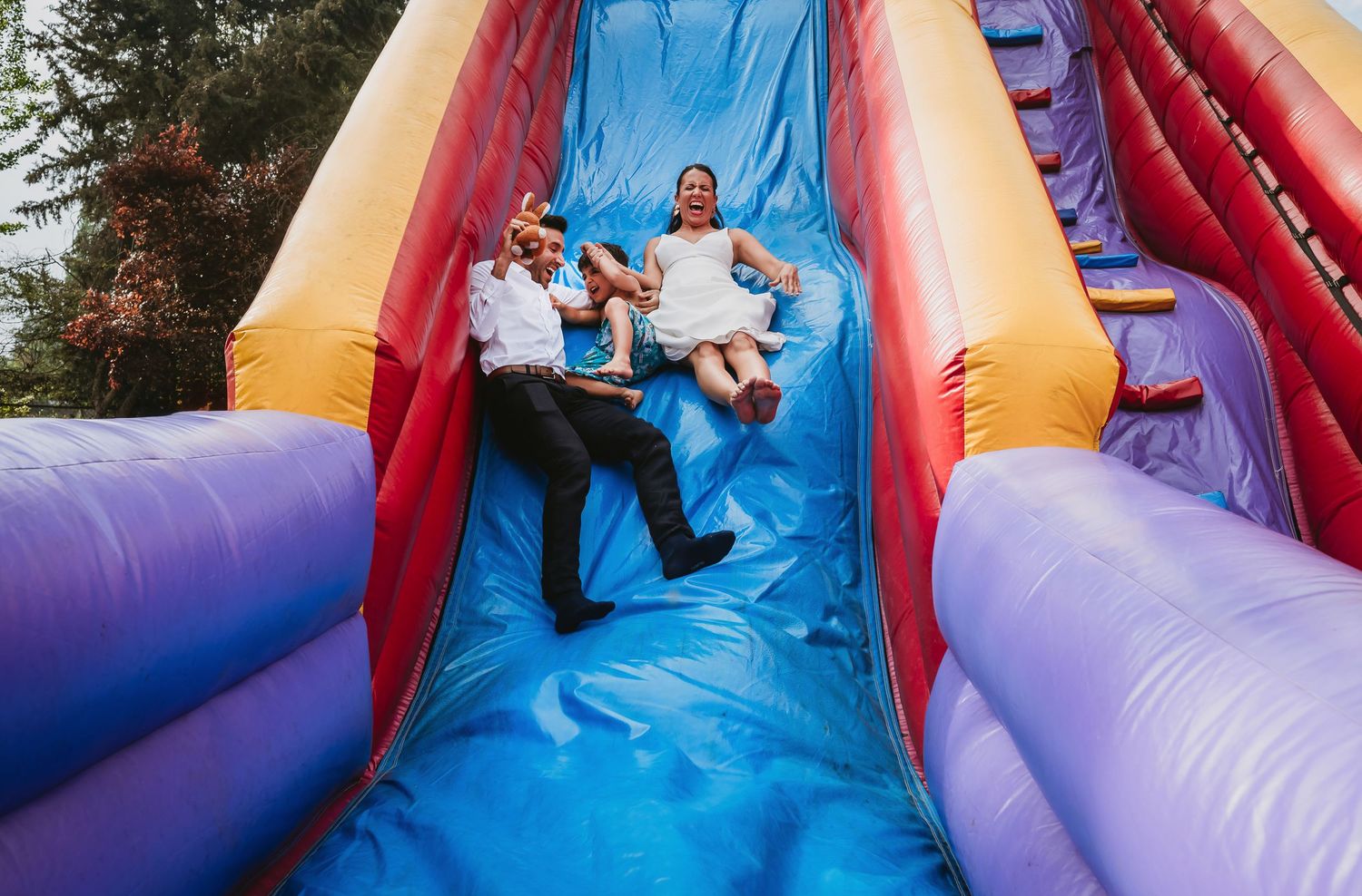 Two people joyfully sliding down a colorful inflatable bounce slide at an outdoor event.