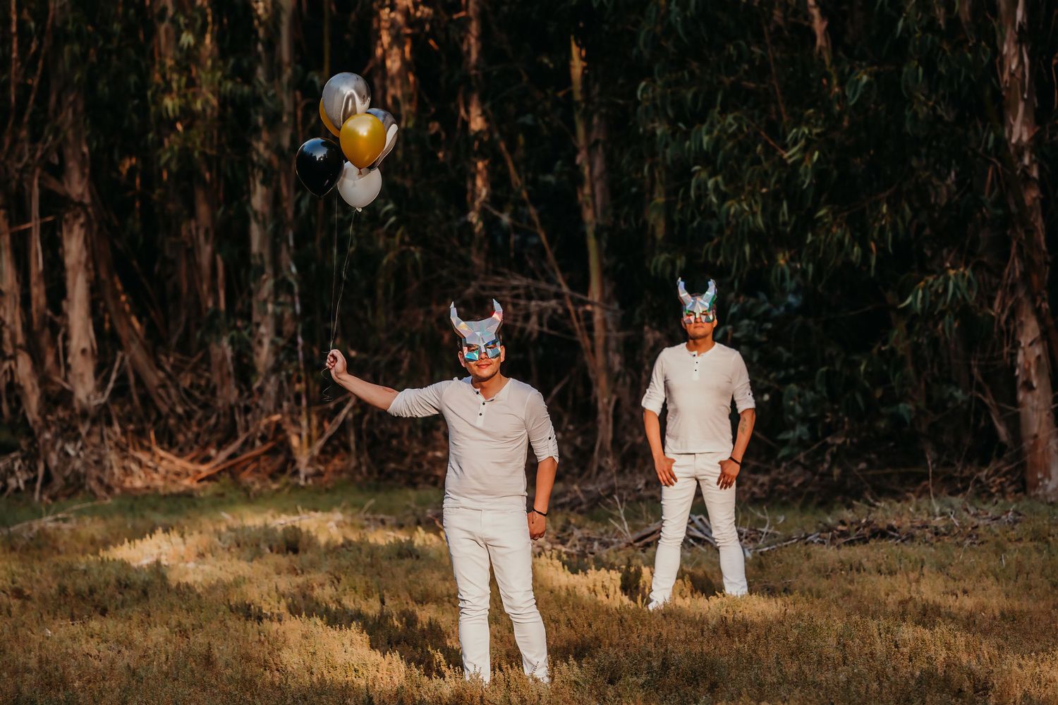 Two men in white outfits and Viking masks stand in a forest holding gold and silver balloons.