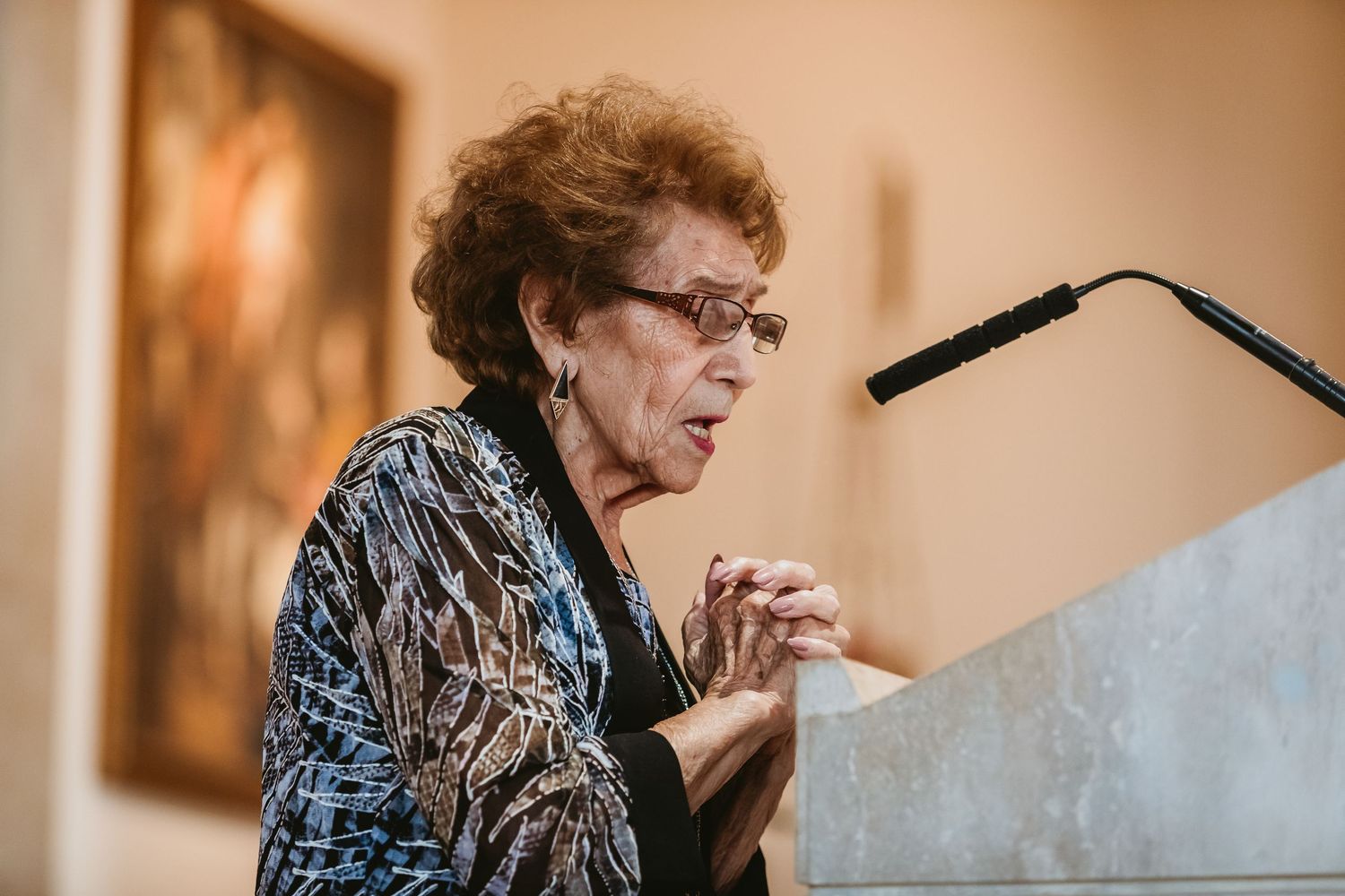 Elderly woman with curly red hair speaking at a podium during a formal event or ceremony.