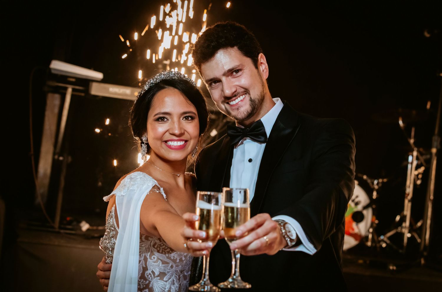 Happy newlyweds clinking champagne glasses outdoors with sparkling fireworks glowing behind them.