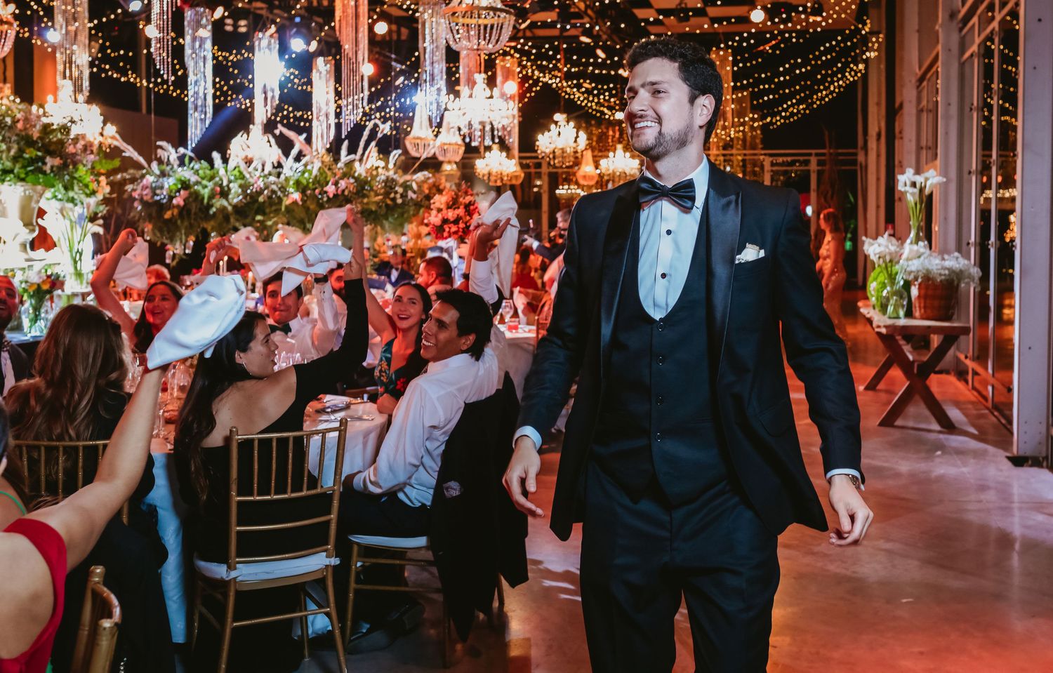 Groom in black tuxedo smiles as he enters elegant floral wedding reception hall with guests.