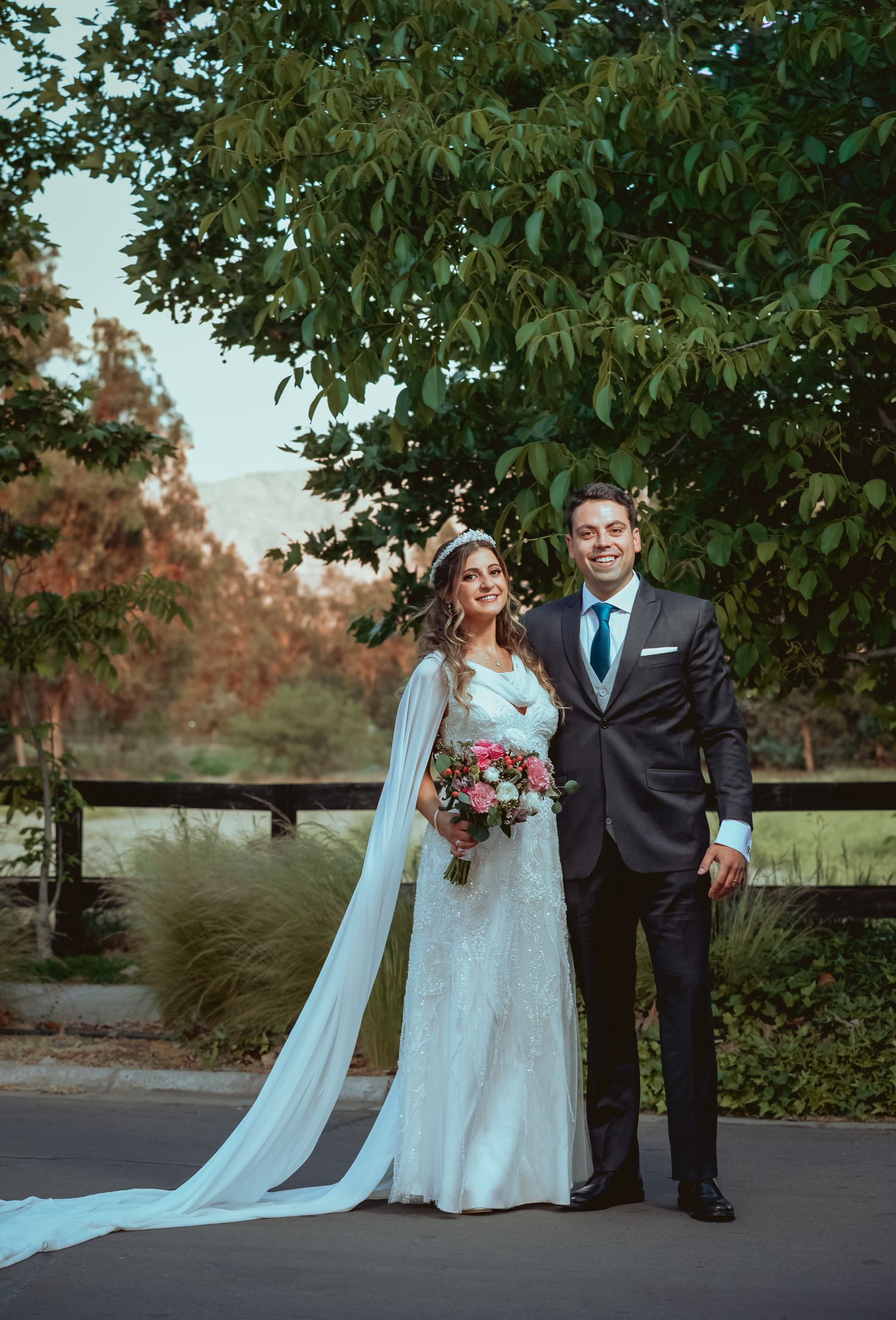 Elegant bride and groom posing together under a large green tree holding a pink and white bridal bouquet.