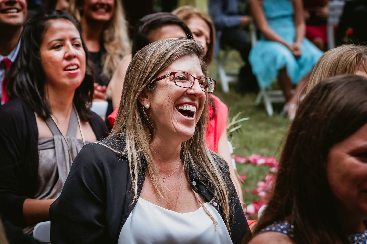 A woman with glasses laughs joyfully while seated among wedding guests outdoors.