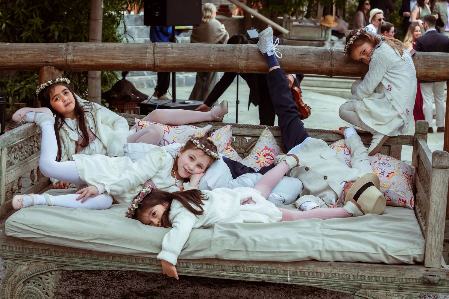Children in white formal attire laughing and lounging on ornate outdoor bench.