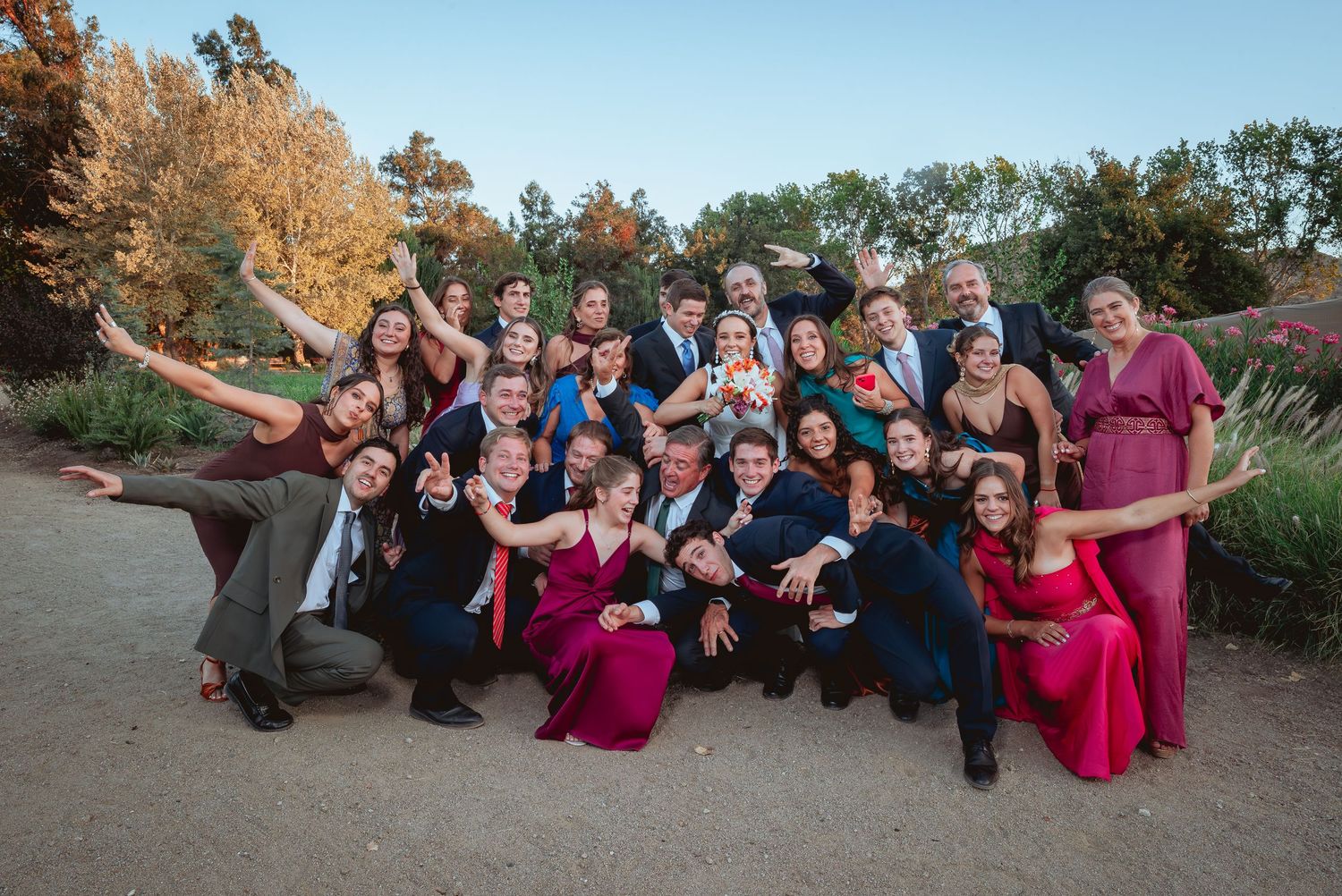 Large wedding party posing playfully outdoors with autumn trees in the background at sunset.