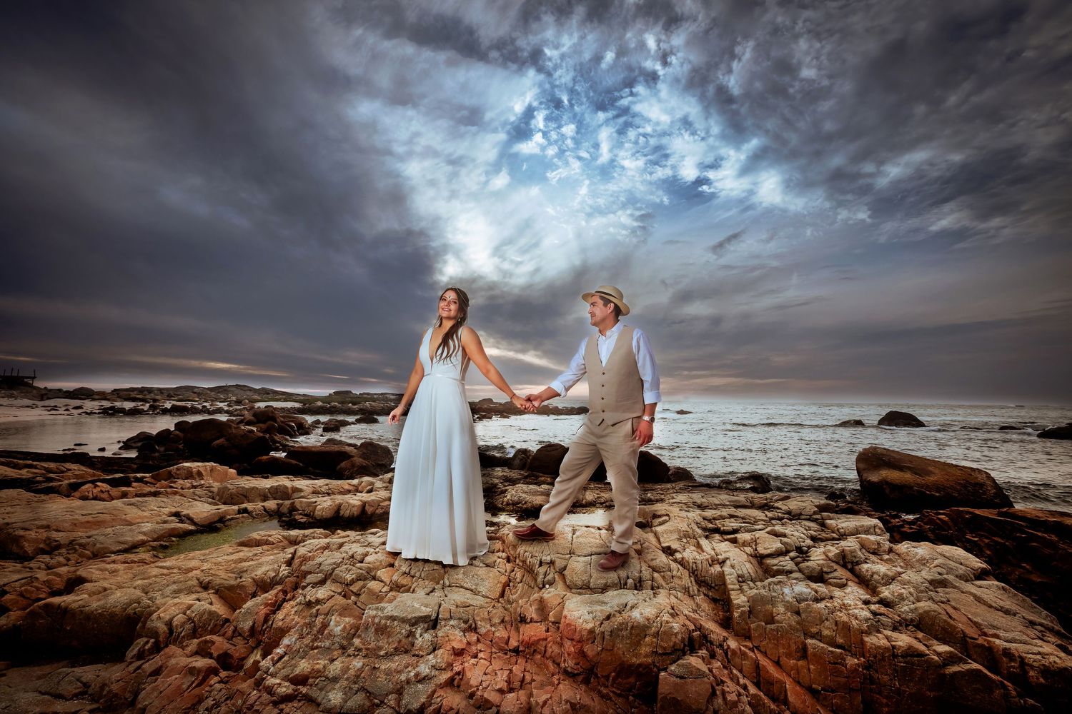 Couple holding hands on rocky beach with dramatic stormy sky at sunset wedding photo.