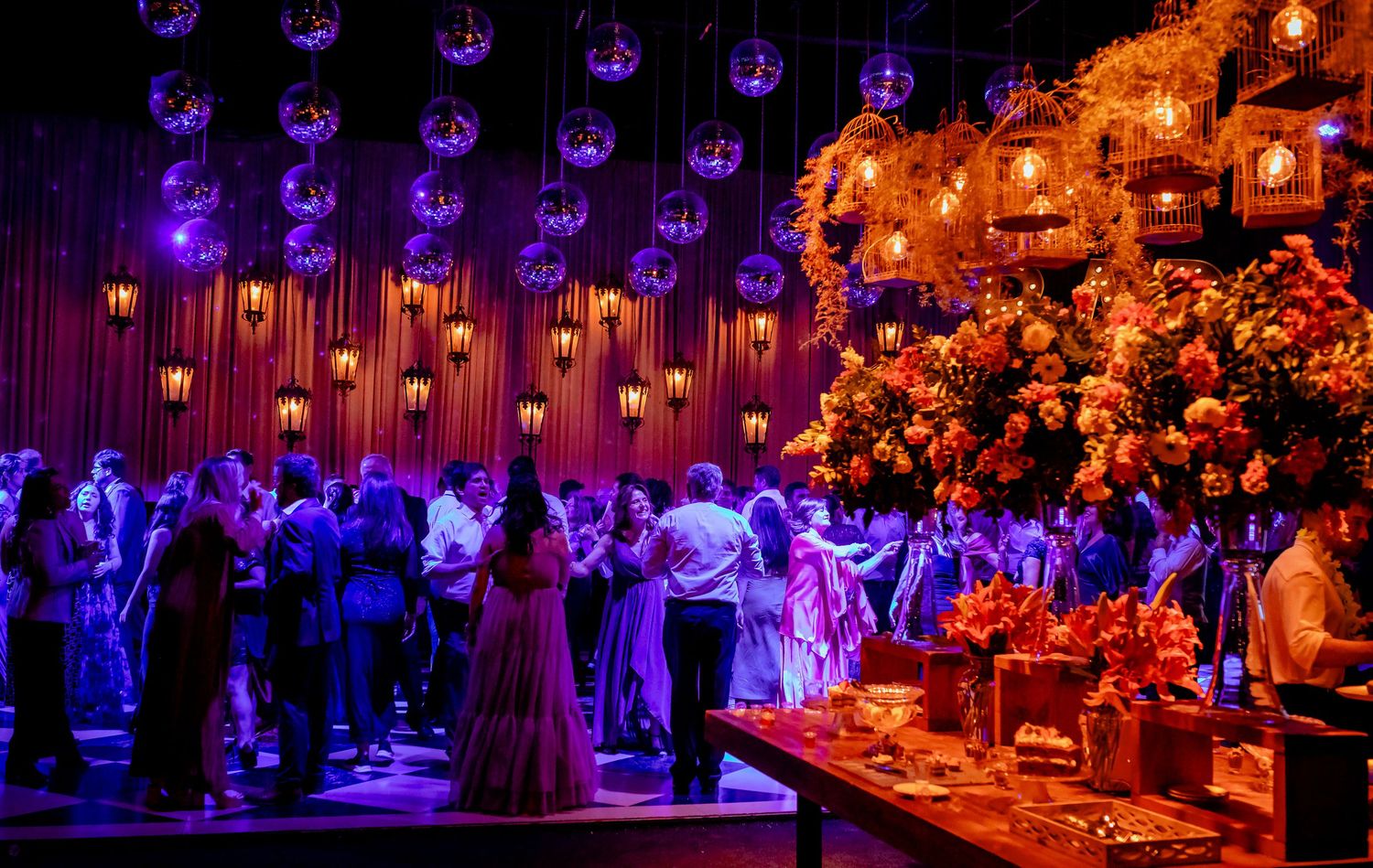 Elegant wedding reception with warm orange floral decor, chandeliers, and guests dancing under glowing pendant lights.