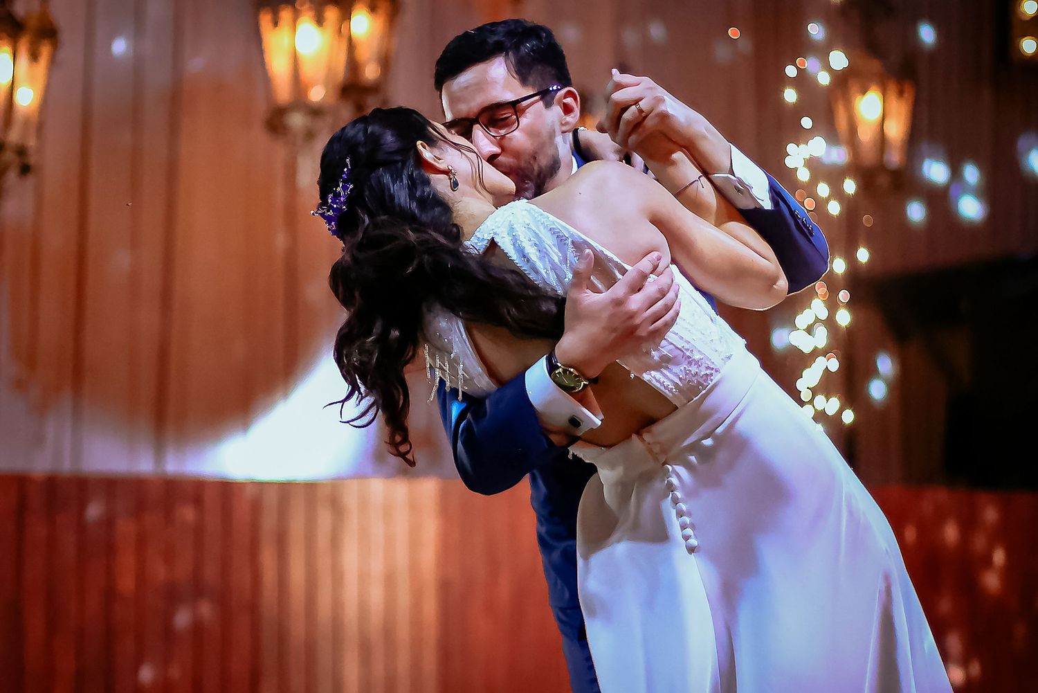 Bride and groom share a romantic first dance kiss at their elegant wedding reception with bokeh lights.