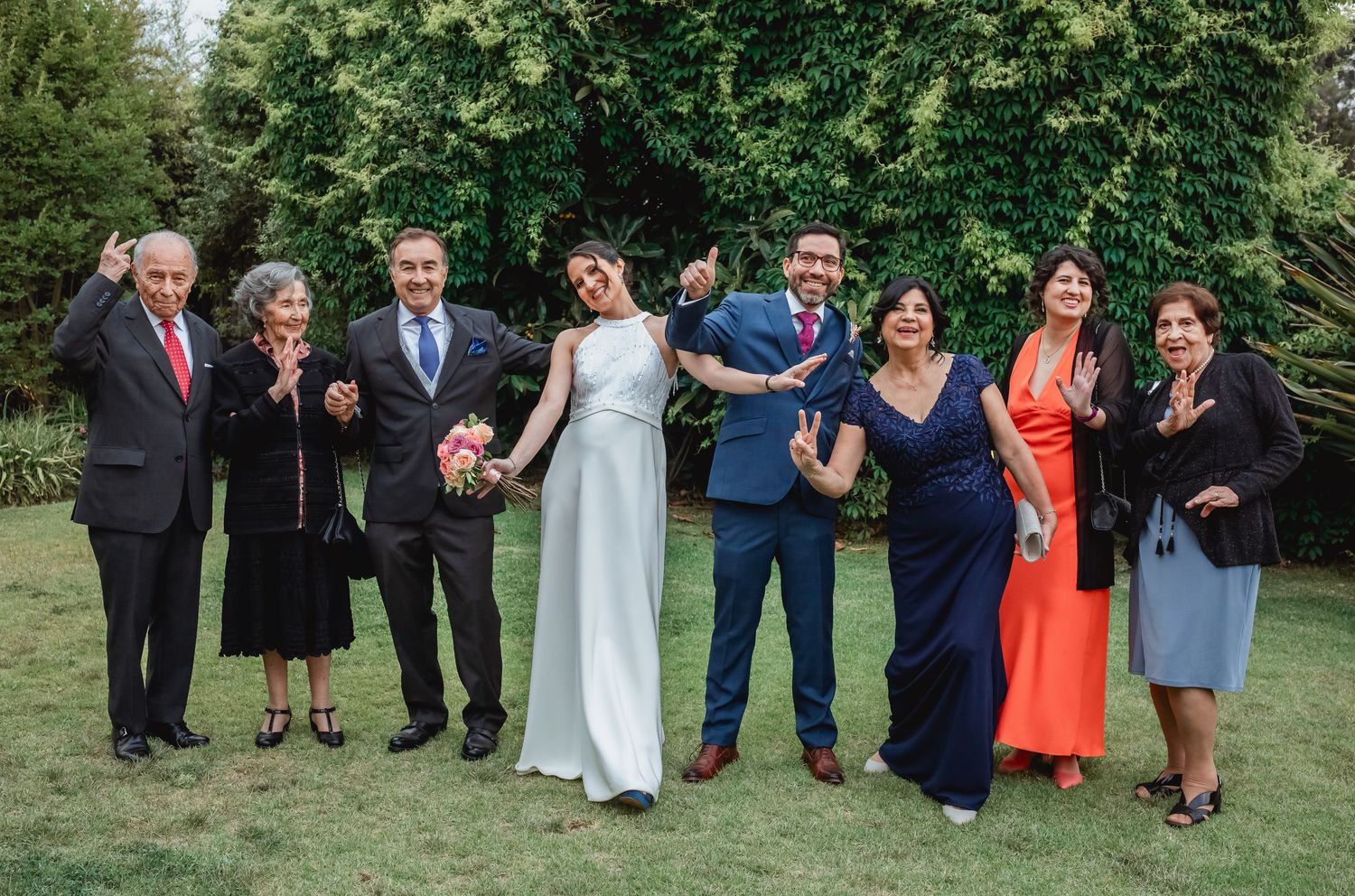 Wedding couple poses with family members on green lawn at outdoor garden reception.