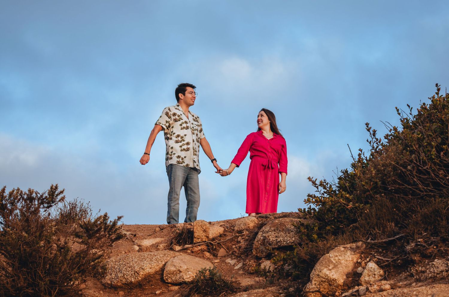 Couple holding hands standing on rocky terrain with blue sky and shrubs in background.