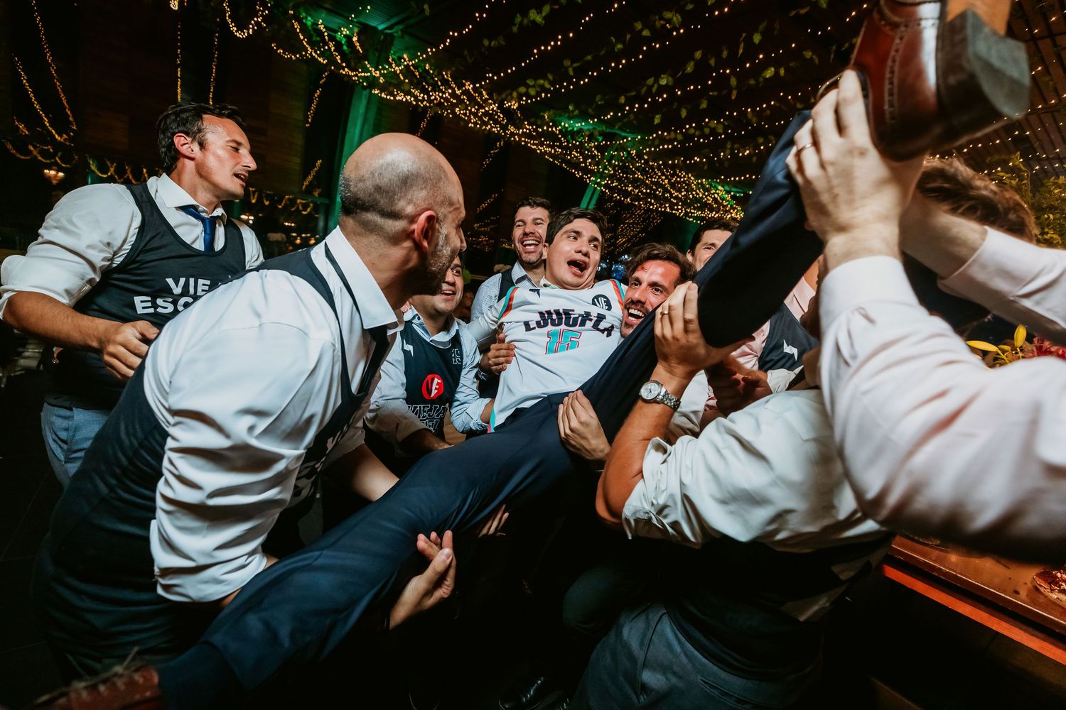 Excited groomsmen in matching jerseys crowd surfing guest at lively outdoor wedding reception.