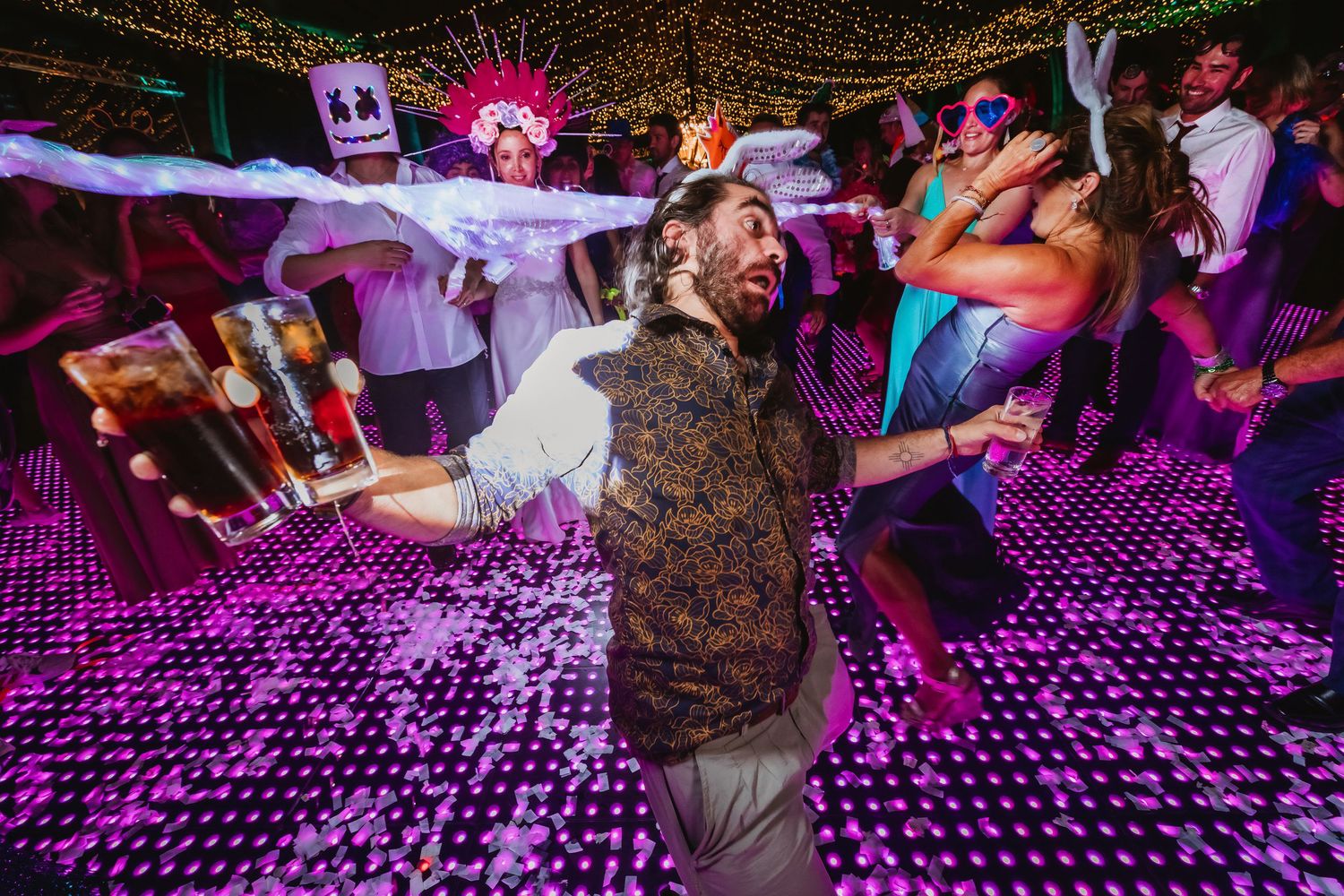 Guest in patterned vest dances energetically on a purple-lit dance floor at a festive wedding reception.