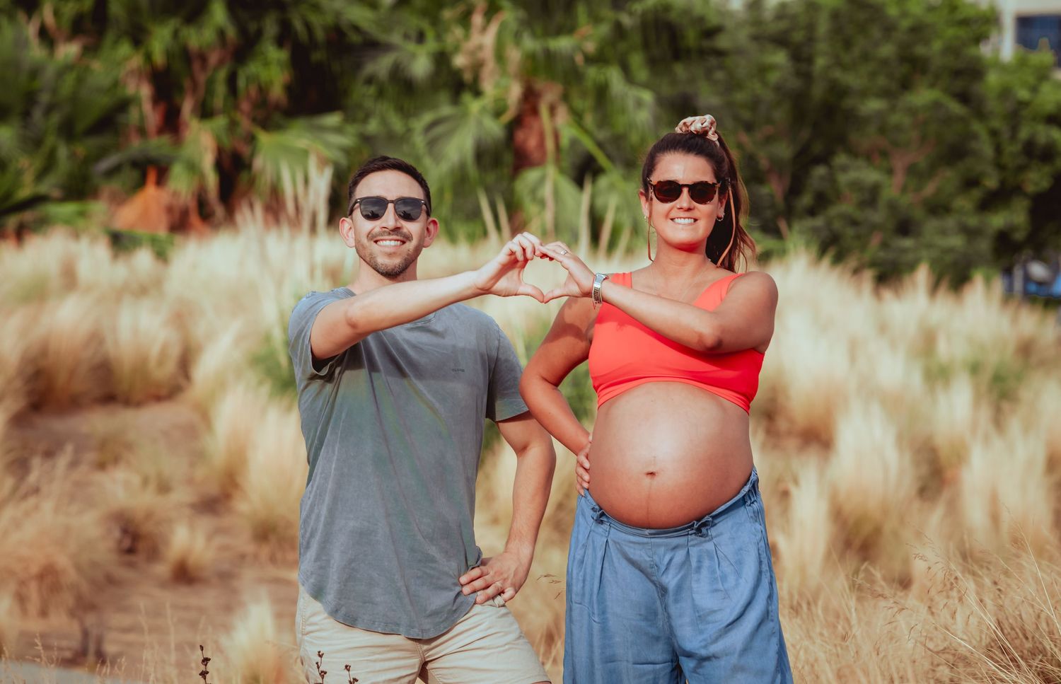 Happy pregnant couple making heart shape with arms outdoors surrounded by golden ornamental grasses.