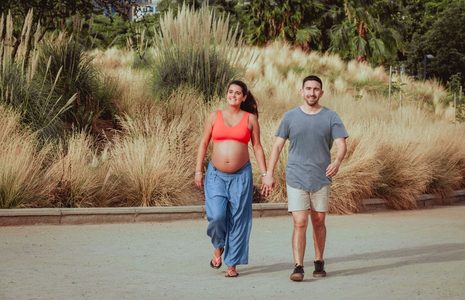 Pregnant couple walking hand in hand on path surrounded by ornamental grasses and tropical palm trees.
