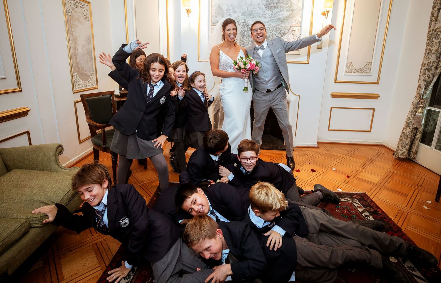 Bride and groom pose with children in school uniforms striking fun poses at an elegant wedding venue.