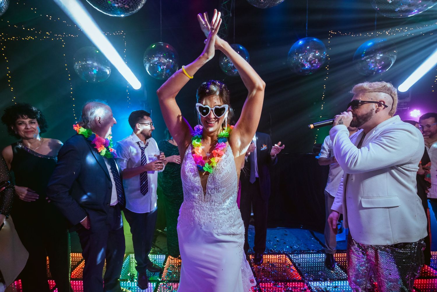 Bride in white dress and novelty sunglasses dancing joyfully at a colorful wedding reception with balloons and live band.