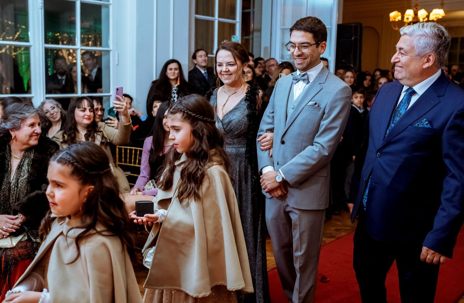 Elegant indoor event with adults and children in formal attire gathered in a decorated hall with red carpet.