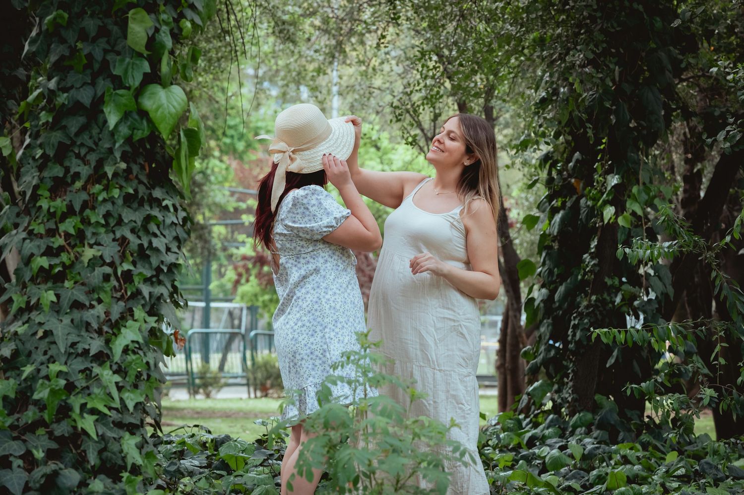 Two women laugh playfully in a lush green garden, one pregnant in white dress and straw hat.