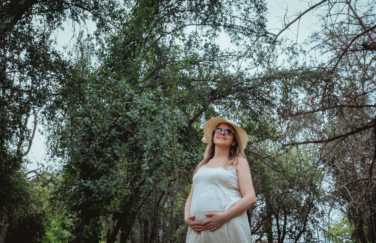 Pregnant woman in white dress and straw hat smiling under green trees outdoors.