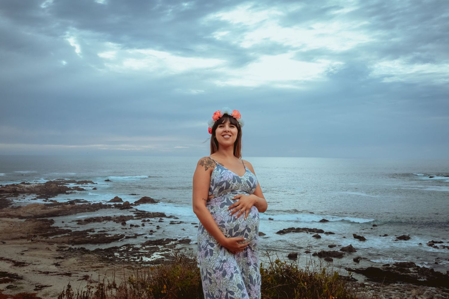 Pregnant woman in floral dress with flower crown smiling by rocky ocean coastline.