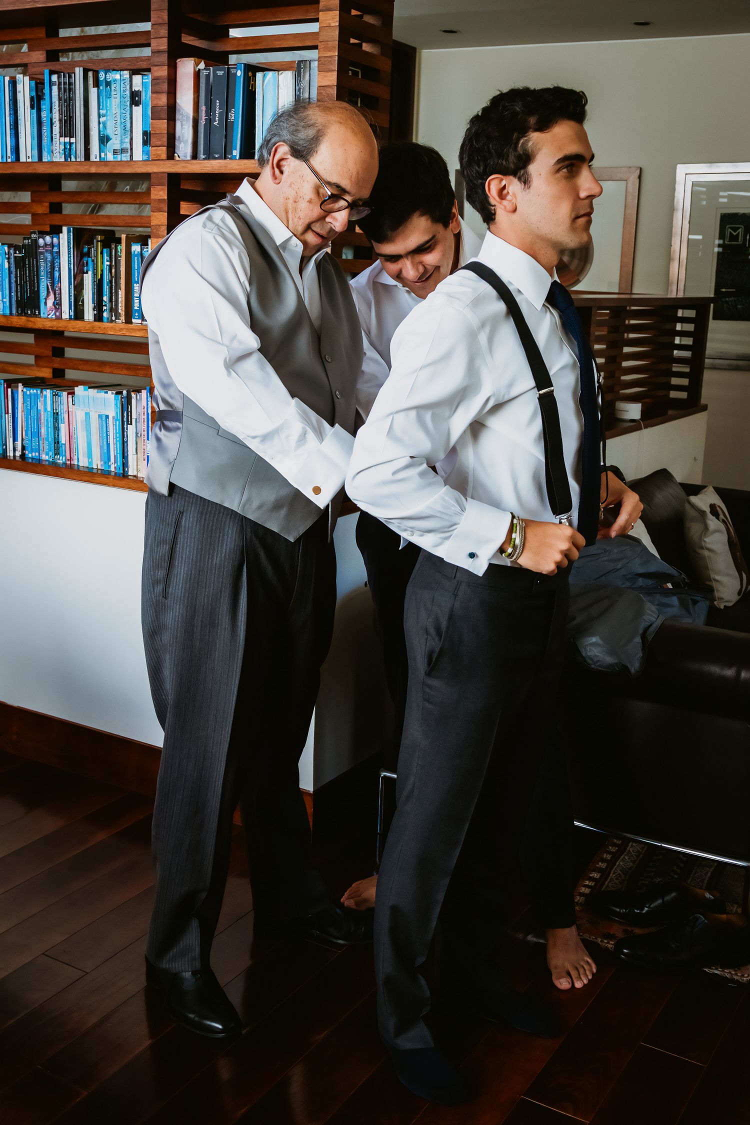 Groom being helped with suspenders by family members in room with wooden bookshelf.