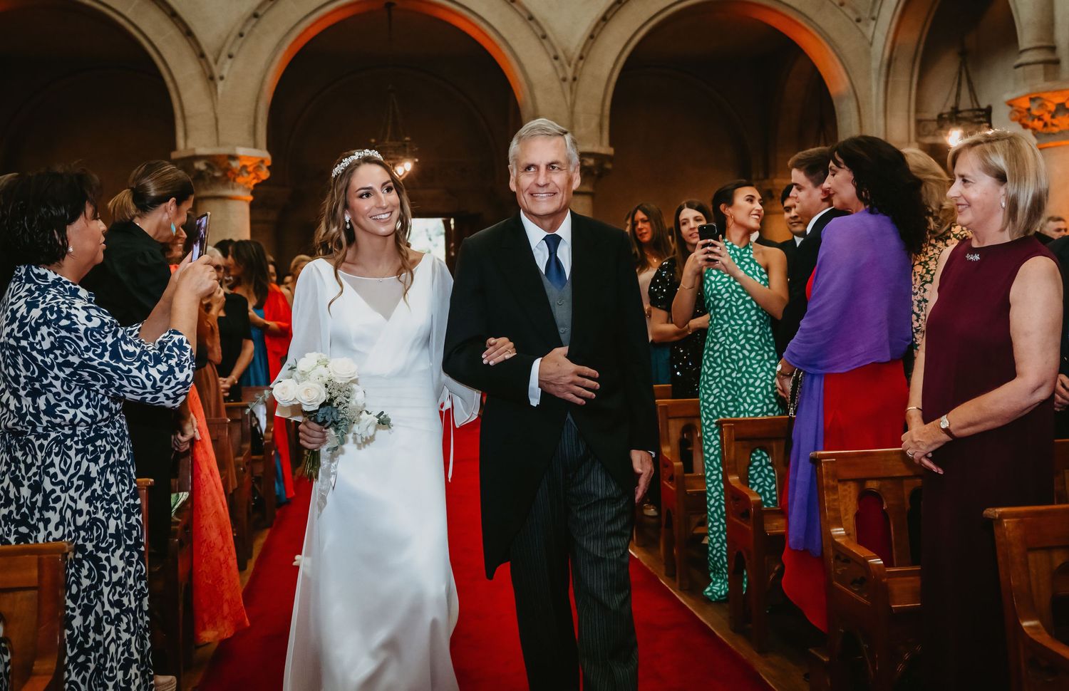 Smiling bride in white gown walking red carpet aisle with escort past applauding church guests.