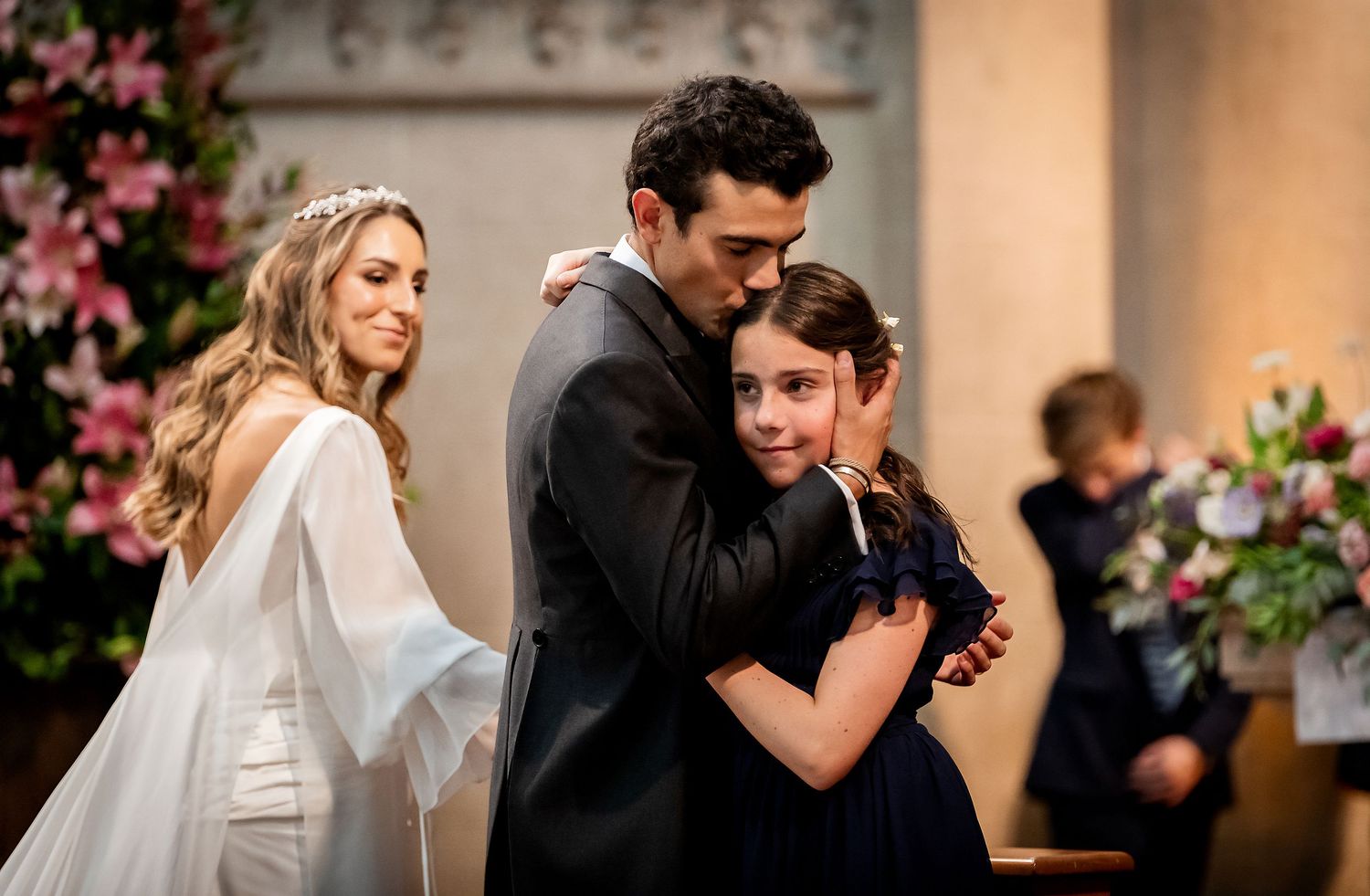 Groom in black suit hugging young girl in navy dress while bride in white stands beside them at church altar.