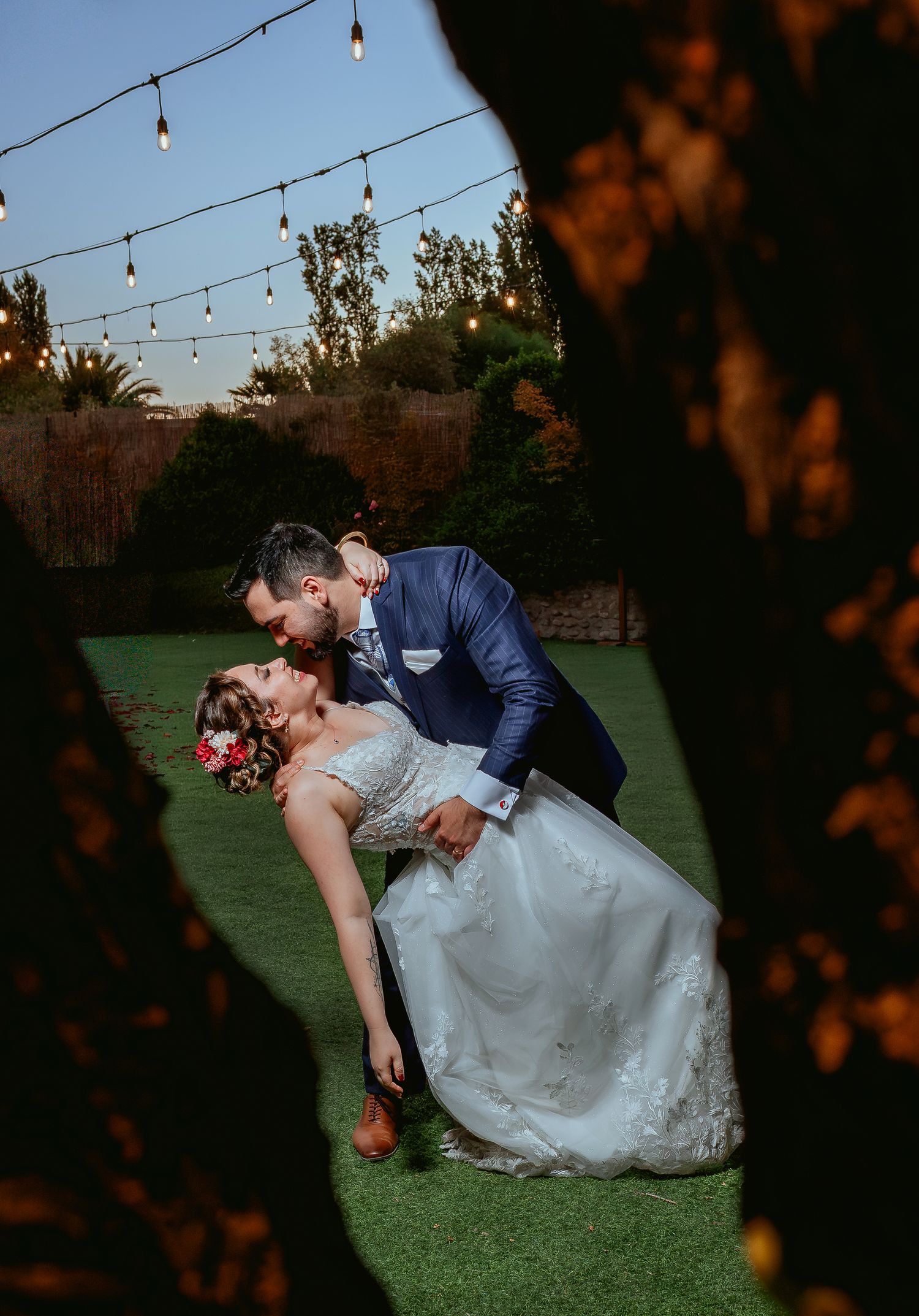 Groom dips bride for a kiss under glowing string lights in outdoor garden wedding.