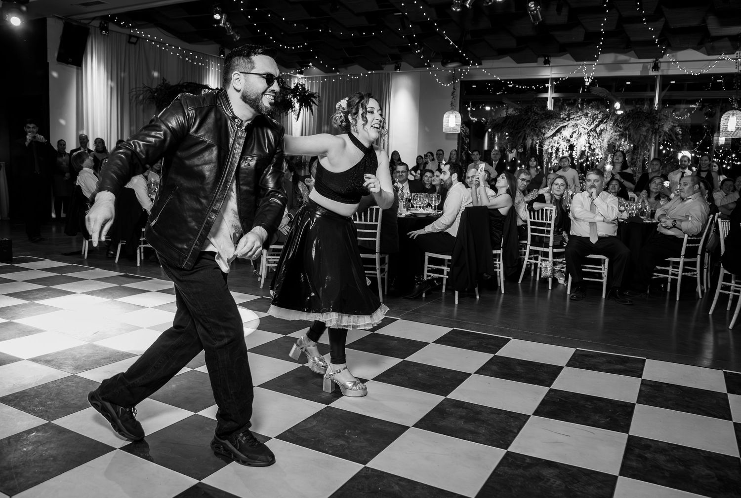 Man in leather jacket dances with woman in black skirt on checkered dance floor at wedding reception.