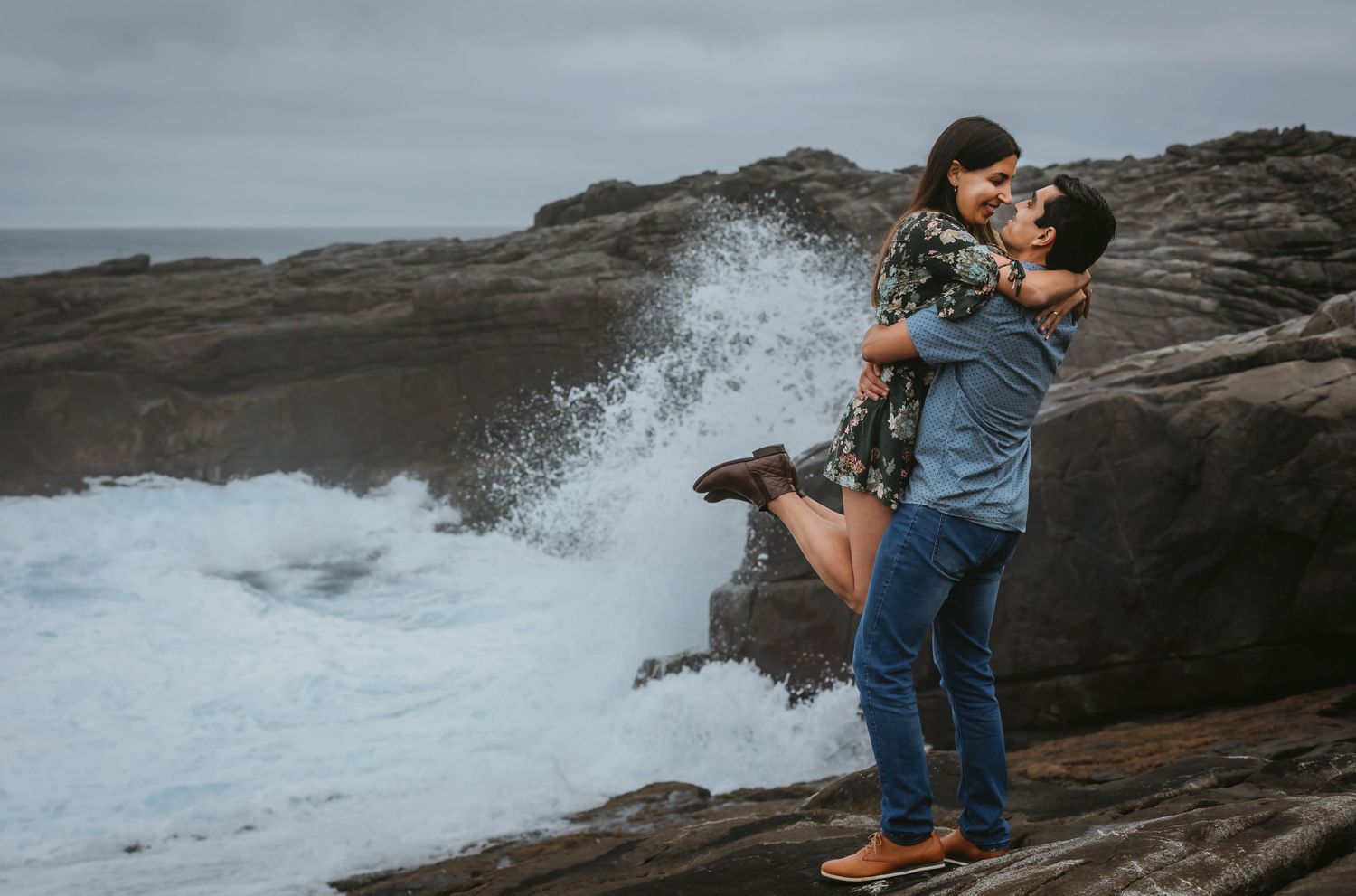 Man lifting woman joyfully on rocky shore as large ocean wave crashes dramatically behind them.