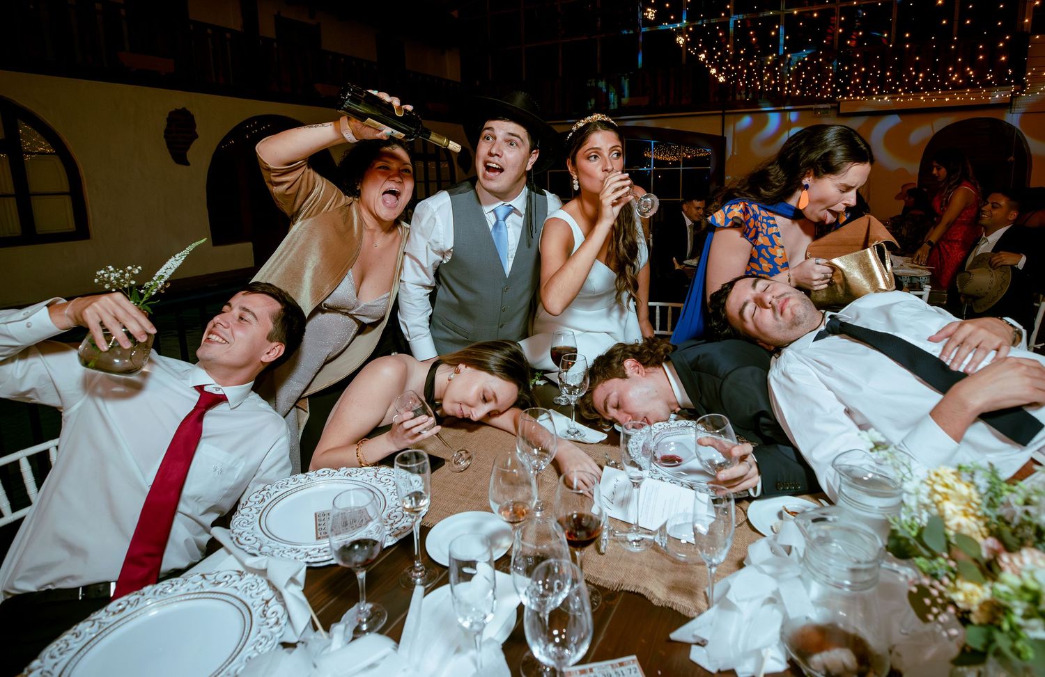 Wedding guests and couple laugh and pose dramatically across decorated banquet table.