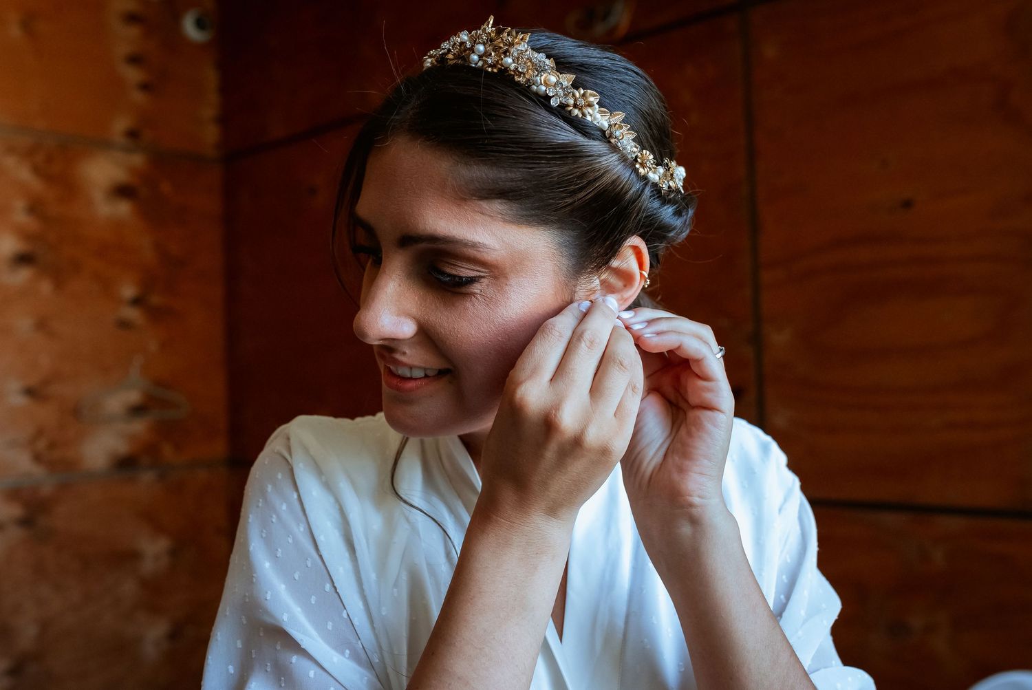 Bride in white robe putting on earrings while wearing an elegant gold floral tiara on her wedding day.