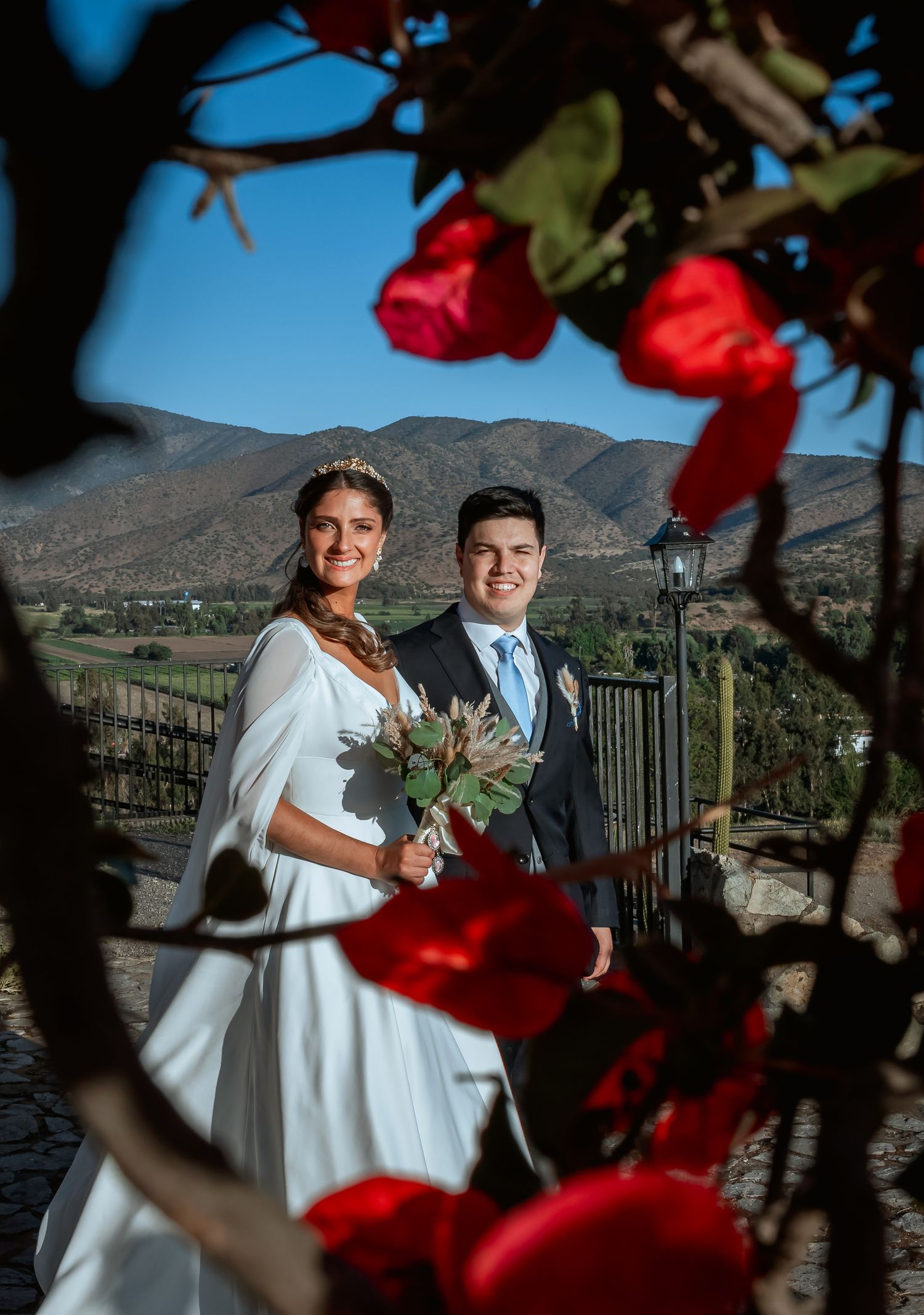 Bride and groom posing romantically framed by red bougainvillea with mountain valley backdrop.