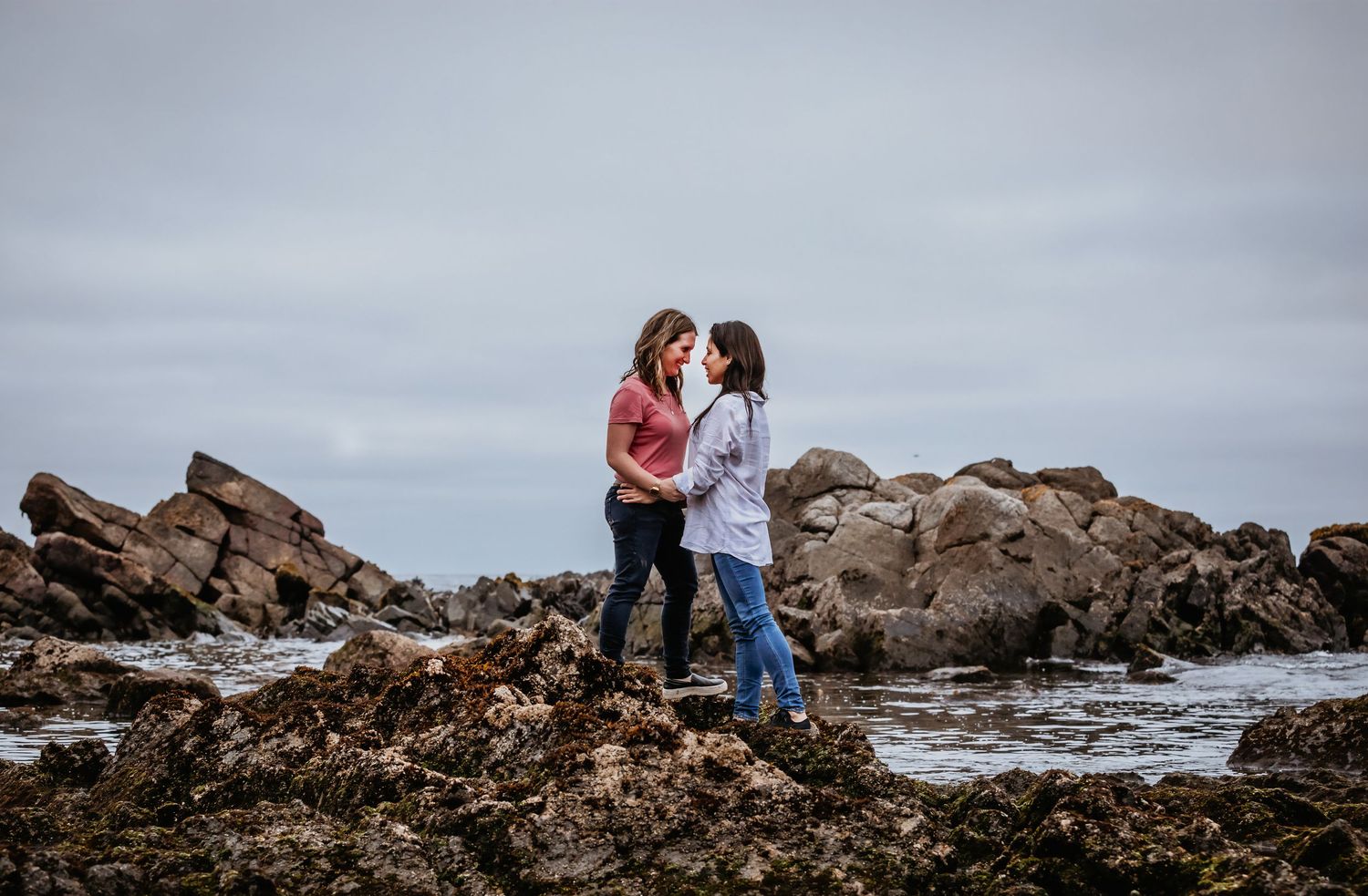Two women hold hands facing each other on rocky shoreline with ocean backdrop.
